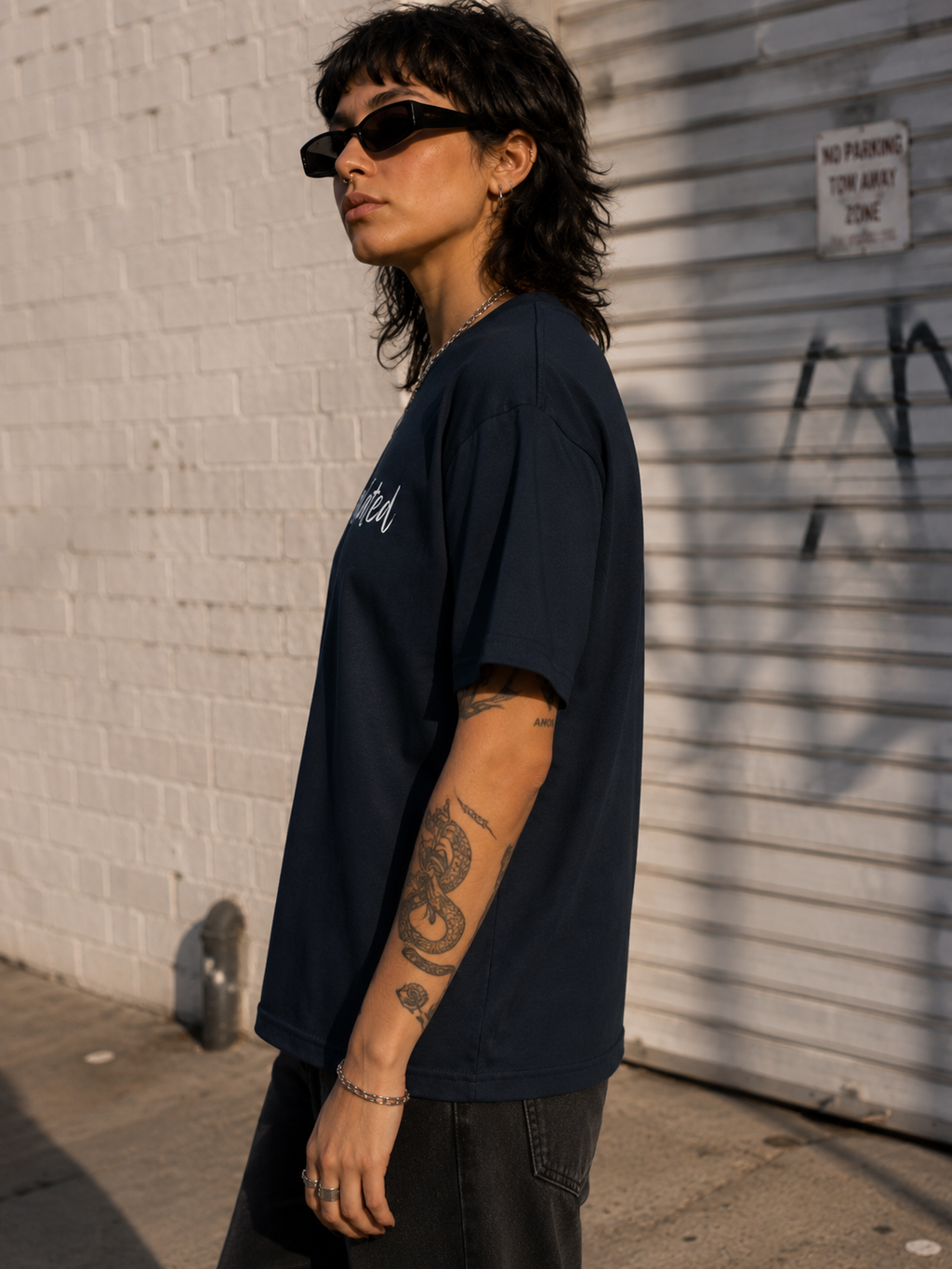 A person wearing an oversized dark navy T-shirt with white script text on the chest, styled with black sunglasses, a silver chain, and black pants, standing outdoors against a white brick wall and metal shutter.
