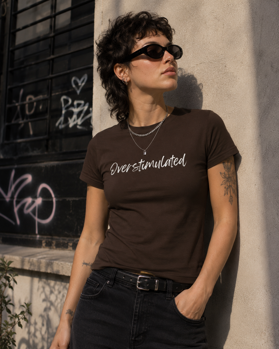 A woman with short curly hair and sunglasses leans against a sunlit concrete wall, wearing a dark brown graphic T-shirt with the phrase 'Overstimulated' in white script across the chest, paired with dark jeans.