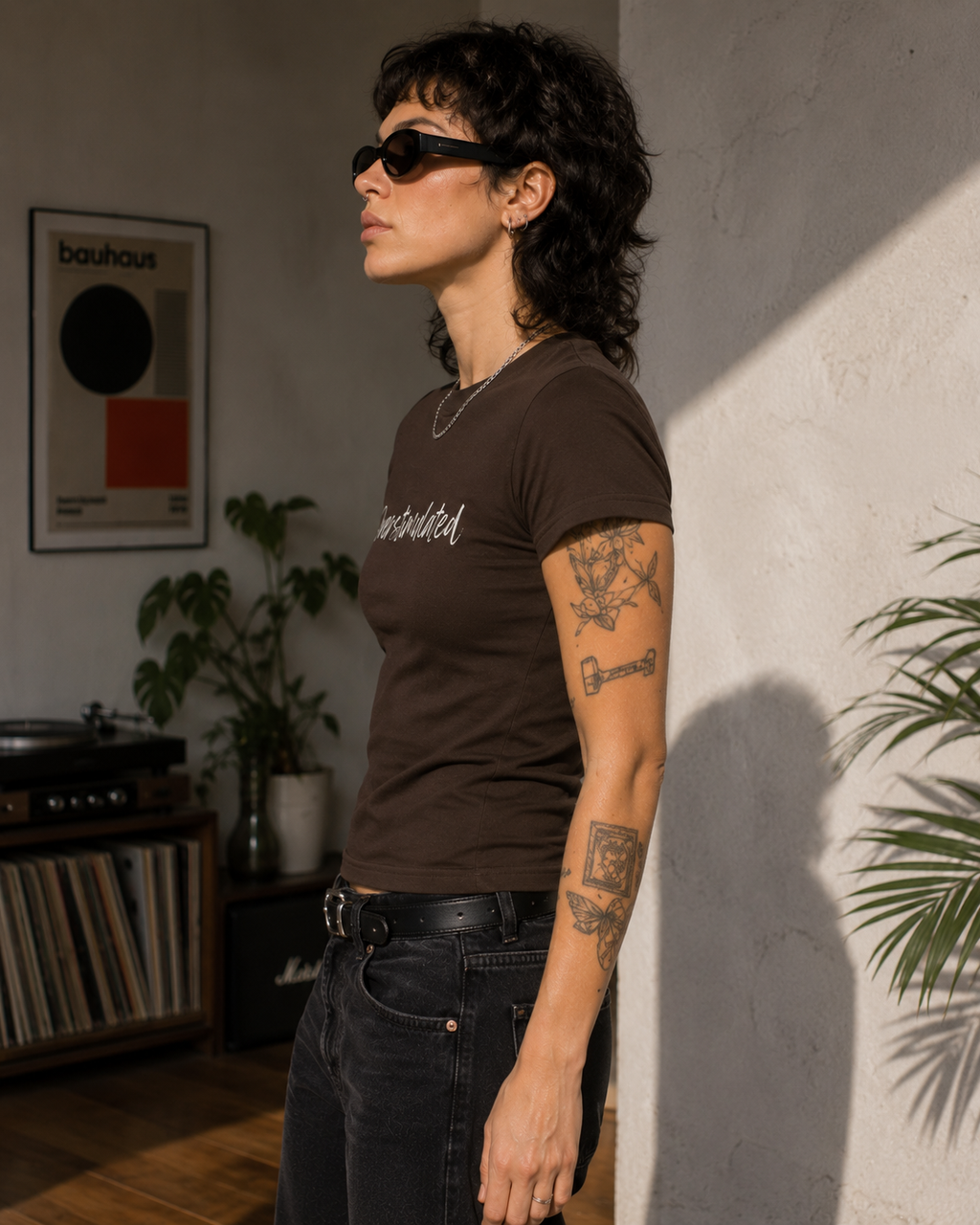 A woman wearing a dark brown t-shirt with white script text and black sunglasses, standing in a sunlit room with a textured wall and plants.