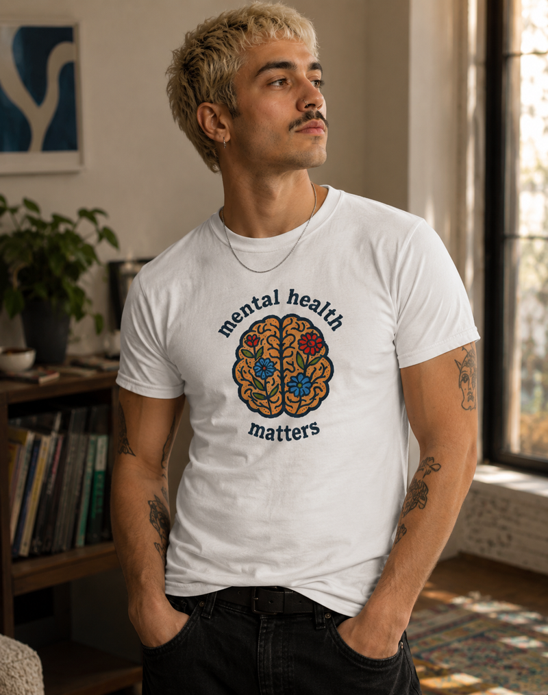 A man wearing a white graphic t-shirt with a colorful brain design and the words 'mental health matters' printed on the front, standing in a cozy indoor setting.