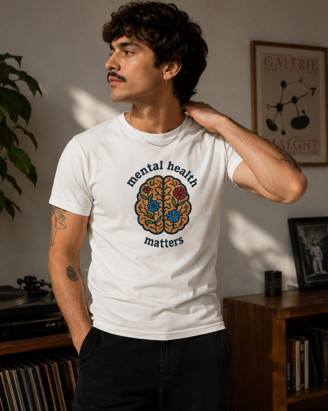 A man wearing a white graphic t-shirt with a colorful brain illustration and the text 'mental health matters' printed on the front, standing in a warmly lit room.