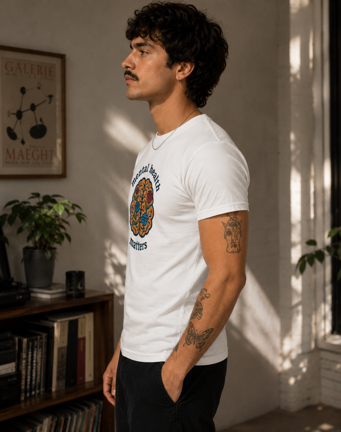 A man wearing a white crewneck t-shirt with a colorful circular graphic on the chest, standing in a sunlit room with a bookshelf and plants in the background.