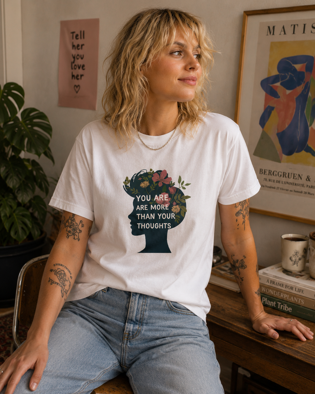 A woman with blonde hair sits in a cozy room wearing a white graphic t-shirt. The t-shirt features a dark teal silhouette of a head filled with colorful flowers and the text 'YOU ARE MORE THAN YOUR THOUGHTS'.