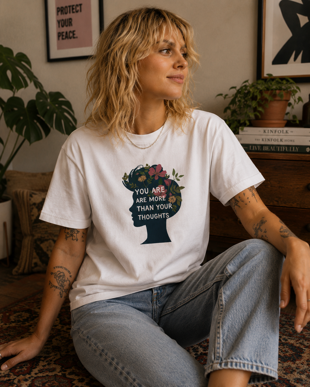 A woman wearing a white graphic t-shirt with a floral silhouette of a head and the text 'YOU ARE MORE THAN YOUR THOUGHTS', paired with light-wash jeans, sitting in a cozy room setting.