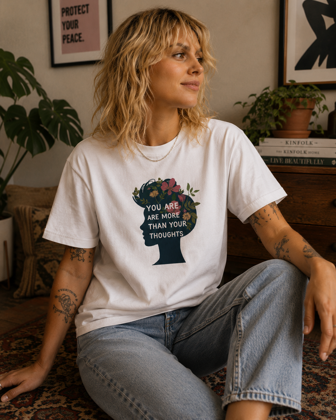 A woman wearing a white graphic t-shirt with a floral silhouette of a head and the text 'YOU ARE MORE THAN YOUR THOUGHTS', paired with light-wash jeans, sitting in a cozy room setting.