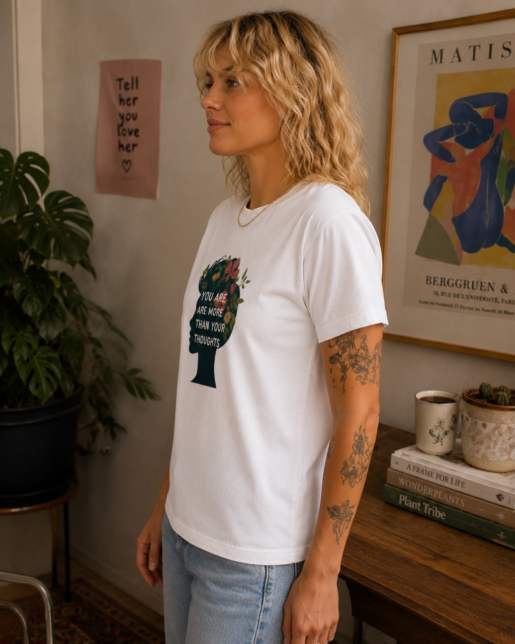 A woman with blonde curly hair wearing a white graphic t-shirt and light blue jeans, standing in a cozy room with a plant, framed art, and a wooden table.