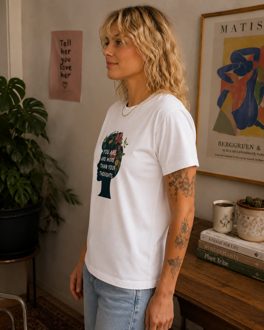 A woman with blonde curly hair wearing a white graphic t-shirt and light blue jeans, standing in a cozy room with a plant, framed art, and a wooden table.
