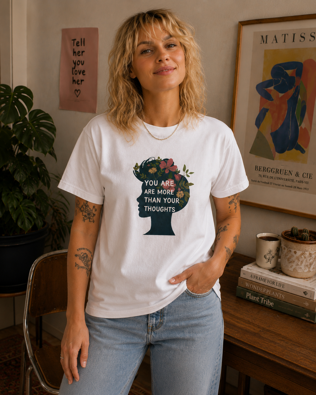 A woman wearing a white graphic t-shirt with a dark teal silhouette of a head filled with flowers and the text 'YOU ARE MORE THAN YOUR THOUGHTS', paired with light blue jeans in a cozy, decorated room.