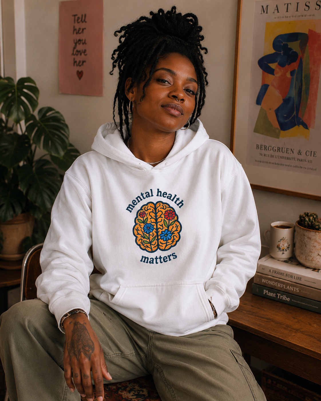 A woman with braided hair sits indoors wearing a white pullover hoodie with a colorful brain graphic and the words 'mental health matters' on the front, paired with olive green pants. The room has framed art, a plant, and books in the background.