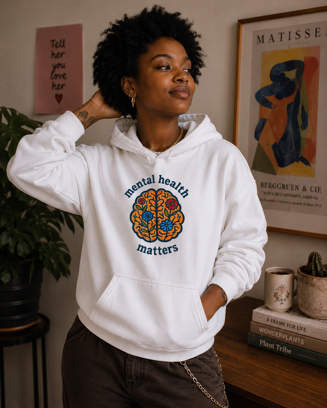 A woman wearing a white pullover hoodie with a colorful brain graphic and the words 'mental health matters' printed on the front, posed in a cozy indoor room setting.