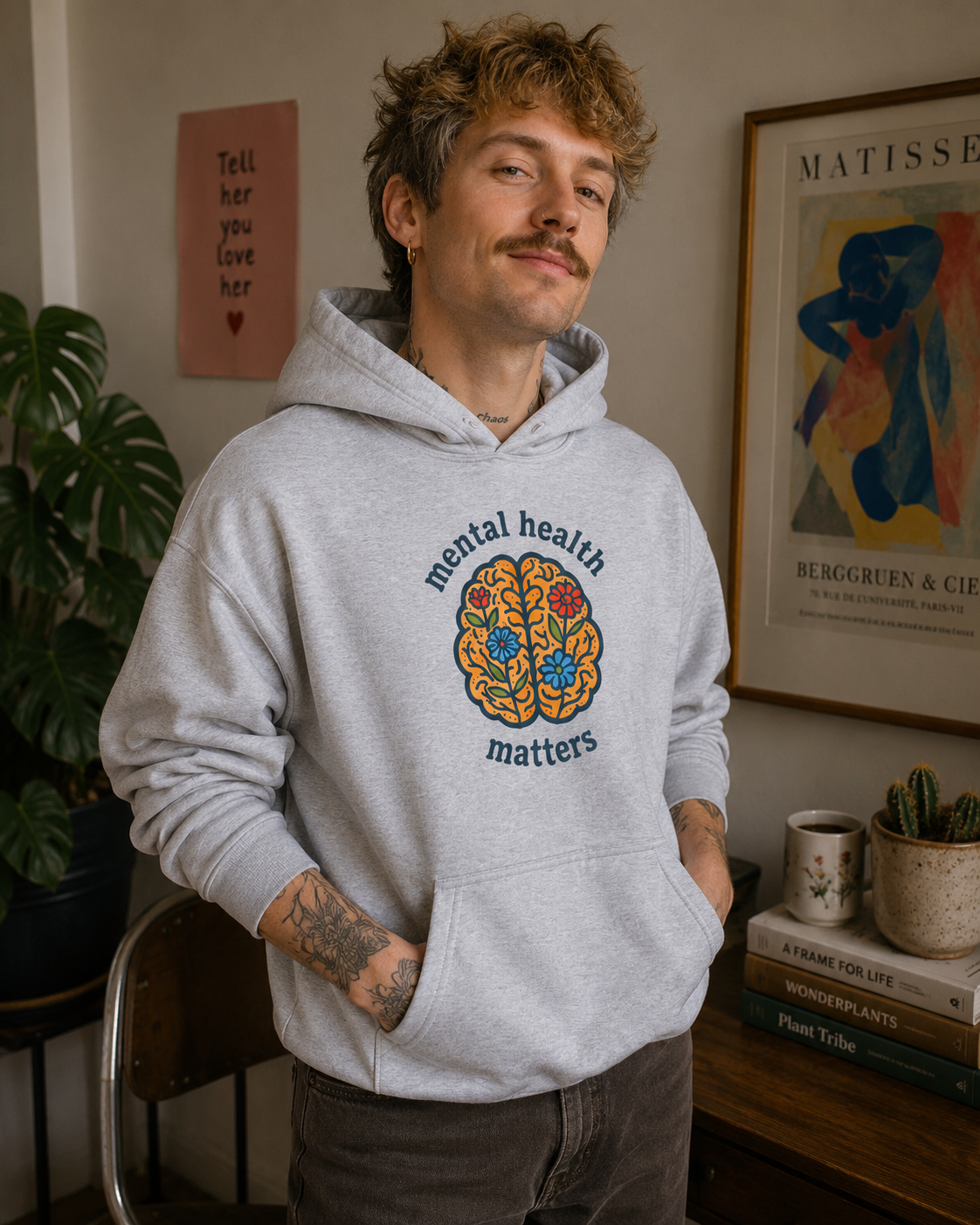 A man wearing a light gray heather hoodie with a colorful brain graphic and the words 'mental health matters' on the front, standing in a cozy indoor room with framed art and plants.