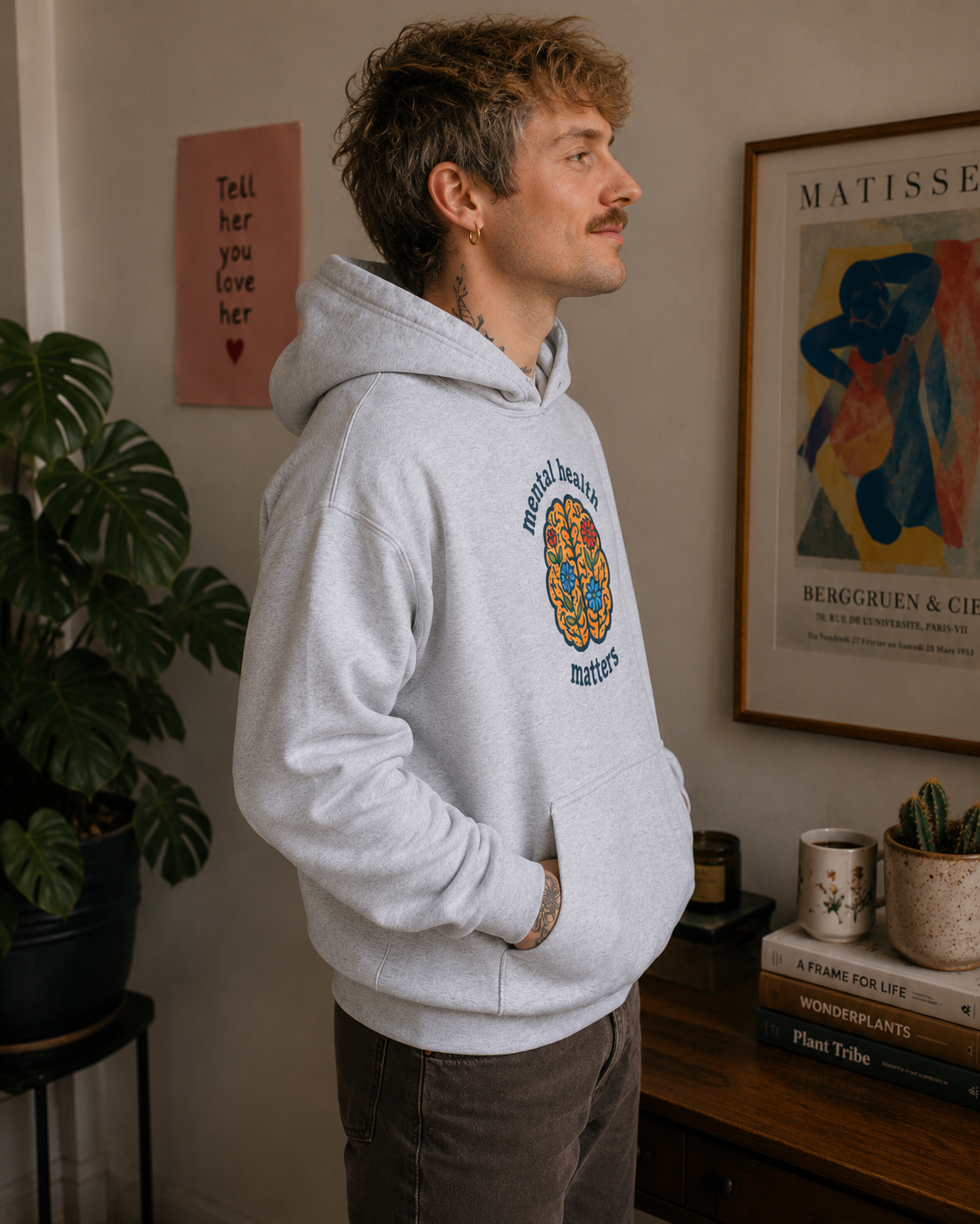 A man wearing a light gray hoodie with a colorful circular graphic on the chest, standing in a room with framed art and a plant in the background.