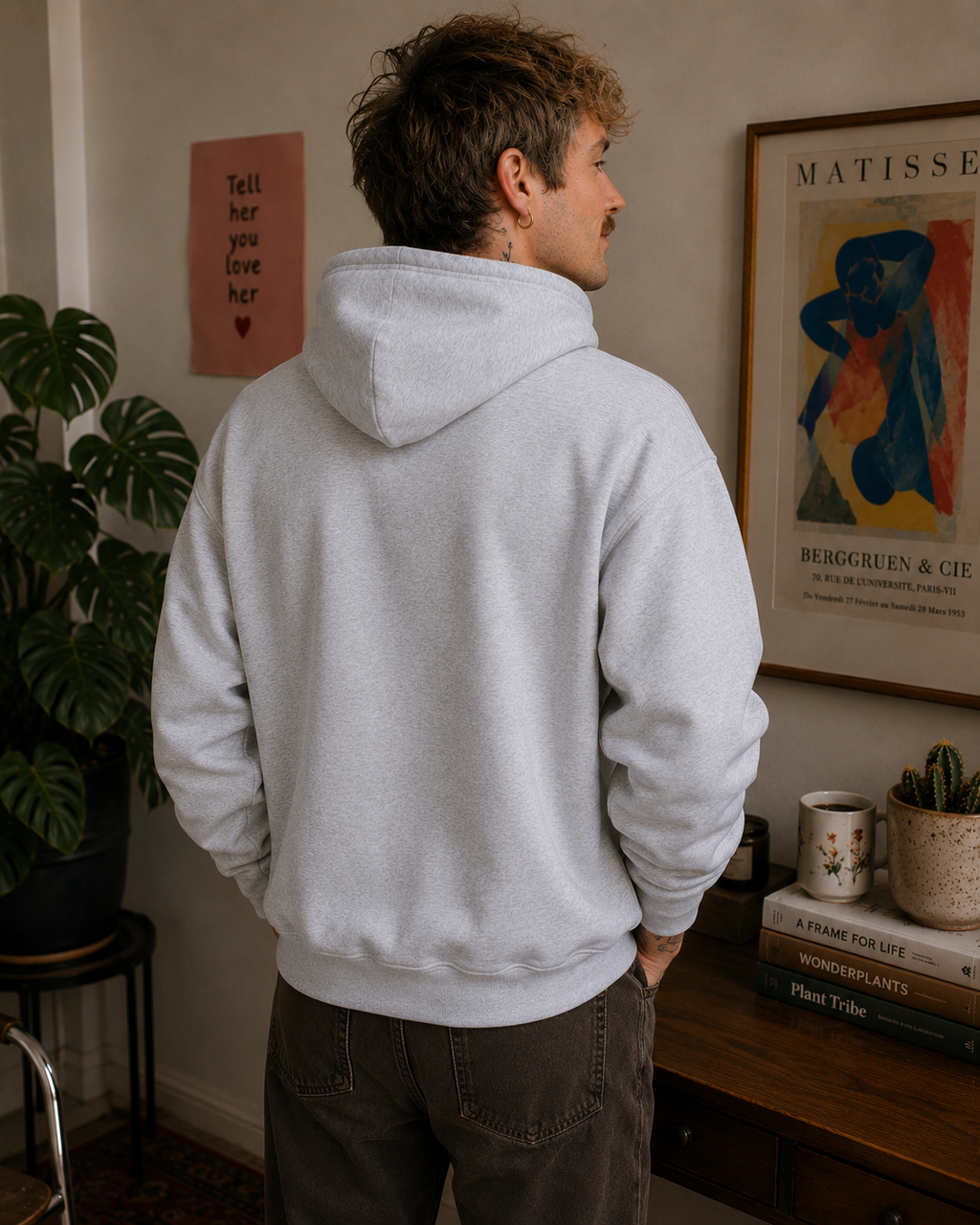A man wearing a light gray heathered hoodie and dark brown jeans, standing in a room with a wooden table, plants, and framed art in the background.
