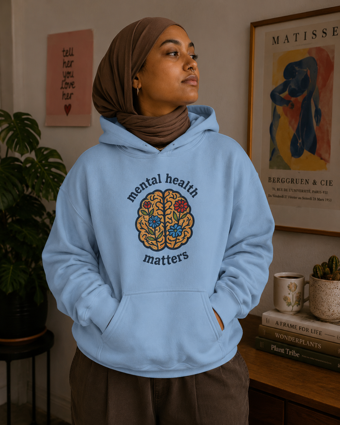 A woman wearing a light blue hoodie with a colorful brain graphic and the words 'mental health matters' stands in a styled indoor room with framed art, a plant, and books.