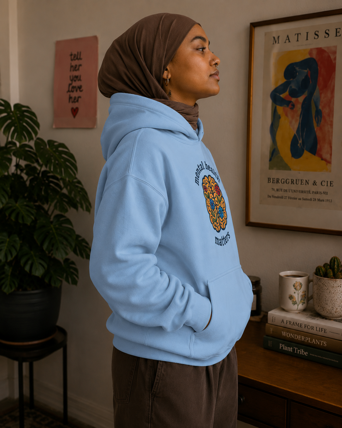 A person wearing a light blue hoodie with a colorful circular graphic on the chest, paired with a brown hijab and brown pants, standing in a decorated indoor room with framed art and a plant.