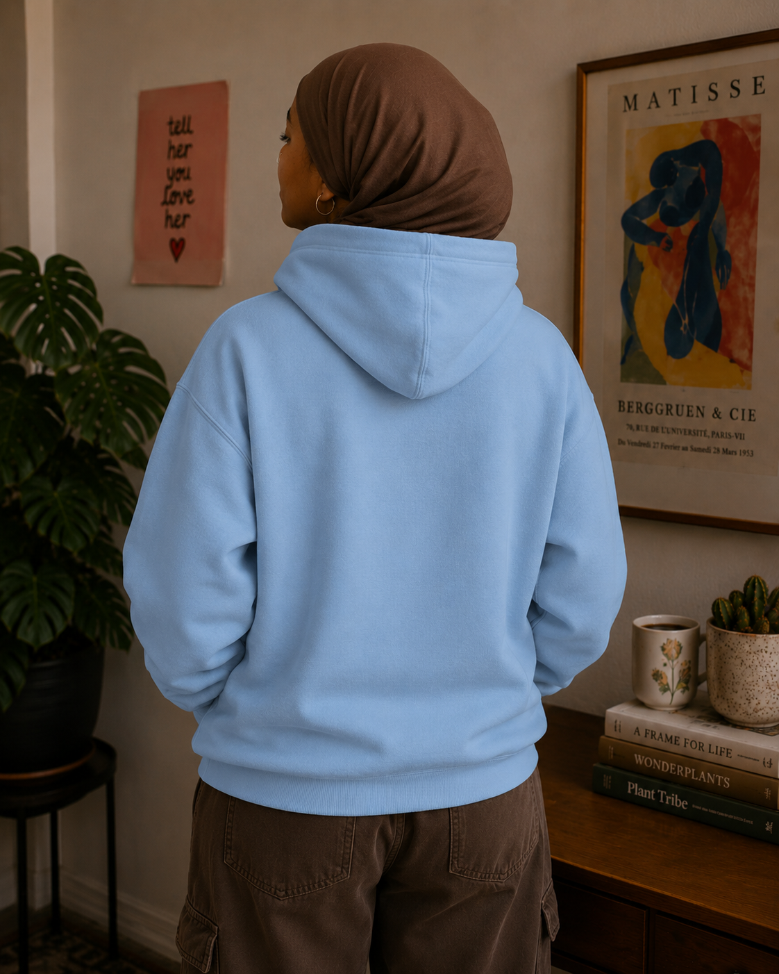 A person wearing a light blue pullover hoodie and brown cargo pants, viewed from behind, standing in a styled indoor room with framed wall art, a plant, and a wooden table.