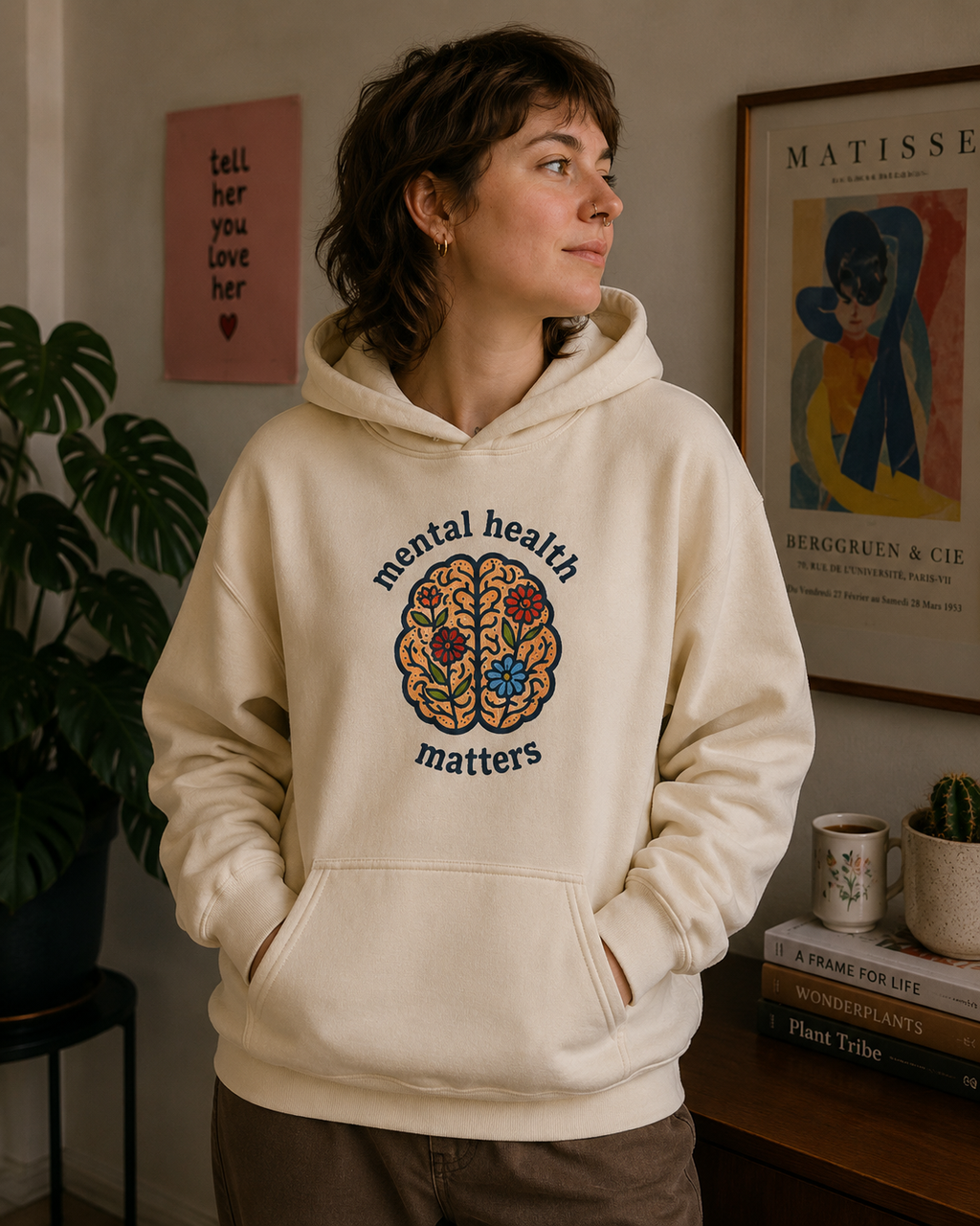 A woman wearing a cream-colored hoodie with a colorful brain graphic and the words 'mental health matters' printed on the front, standing in a cozy indoor setting with framed art and a plant in the background.