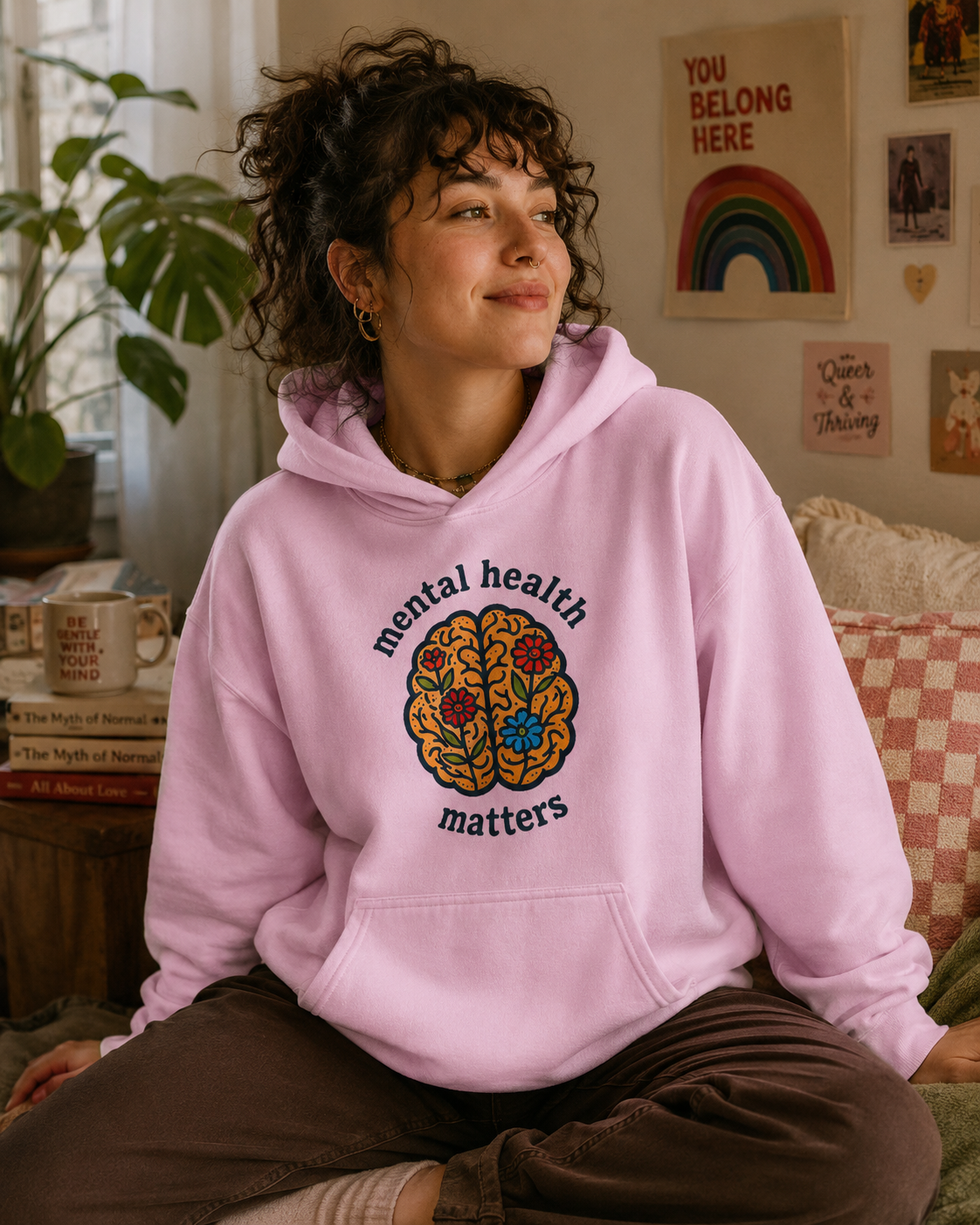 A woman with curly dark hair sits on a couch wearing a light pink hoodie with a colorful brain graphic and the words 'mental health matters'. The background shows a cozy room with posters, a plant, and home decor.