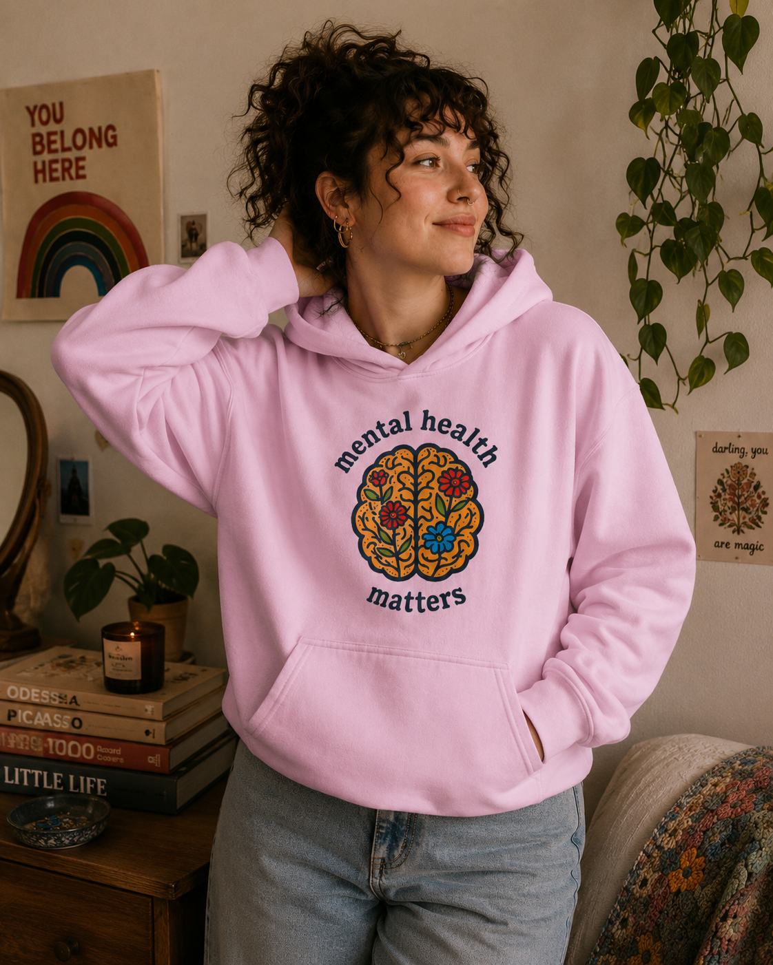 A woman wearing a light pink hoodie with a colorful brain graphic and the words 'mental health matters' on the front, standing in a cozy room setting.