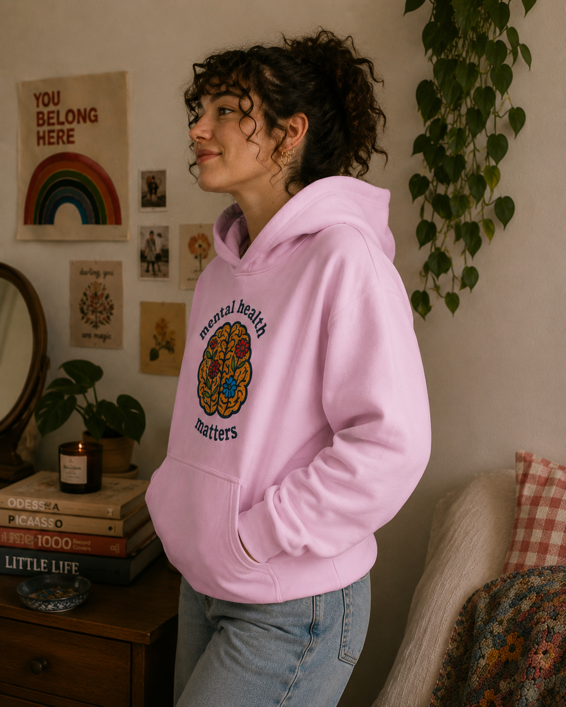 A woman wearing a light pink hoodie with a colorful brain graphic and the words 'mental health matters' stands in a cozy room setting with plants, books, and wall art.