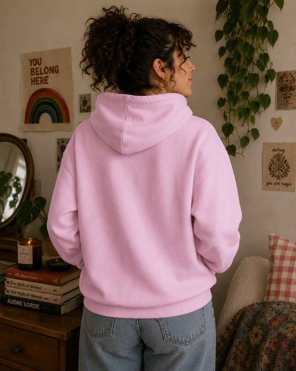 A person wearing a light pink pullover hoodie and light blue jeans, shown from the back in a cozy, decorated room with plants, books, and wall art.