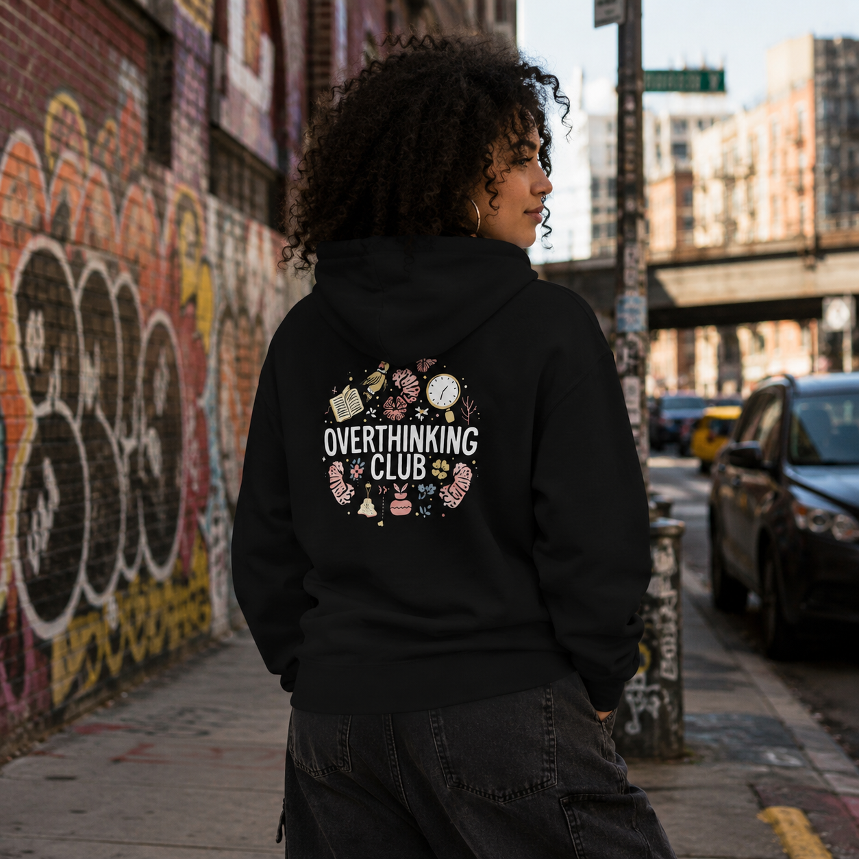 A person standing on a city sidewalk wearing a black hoodie with a graphic on the back that reads 'OVERTHINKING CLUB' surrounded by small illustrated objects. The background shows a graffiti-covered brick wall, urban buildings, and parked cars.