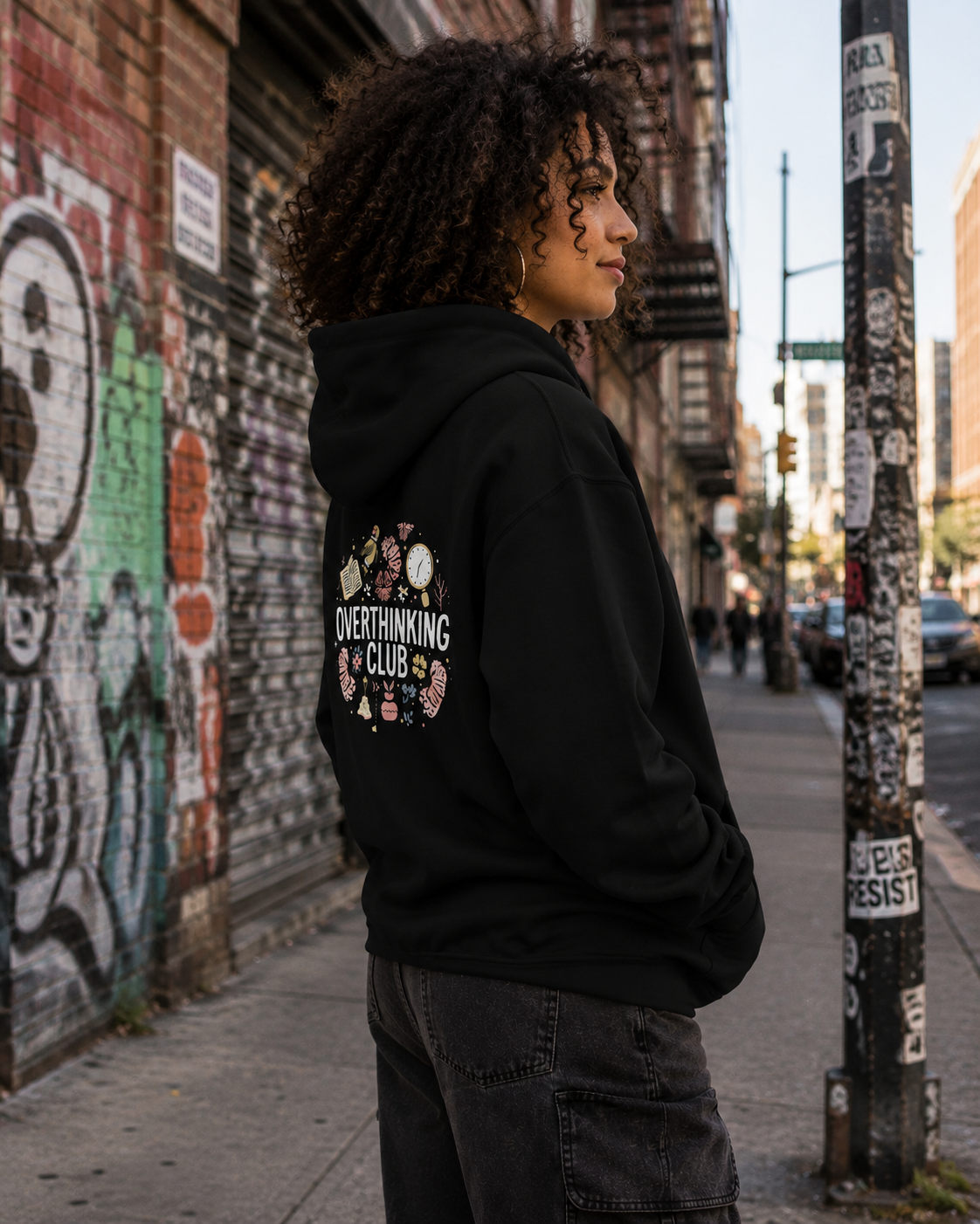 A woman with curly dark hair stands on a city sidewalk wearing a black hoodie with a circular graphic on the back that reads 'OVERTHINKING CLUB' surrounded by small illustrations. The background shows a graffiti-covered brick wall, a street pole with stickers, and an urban street scene.
