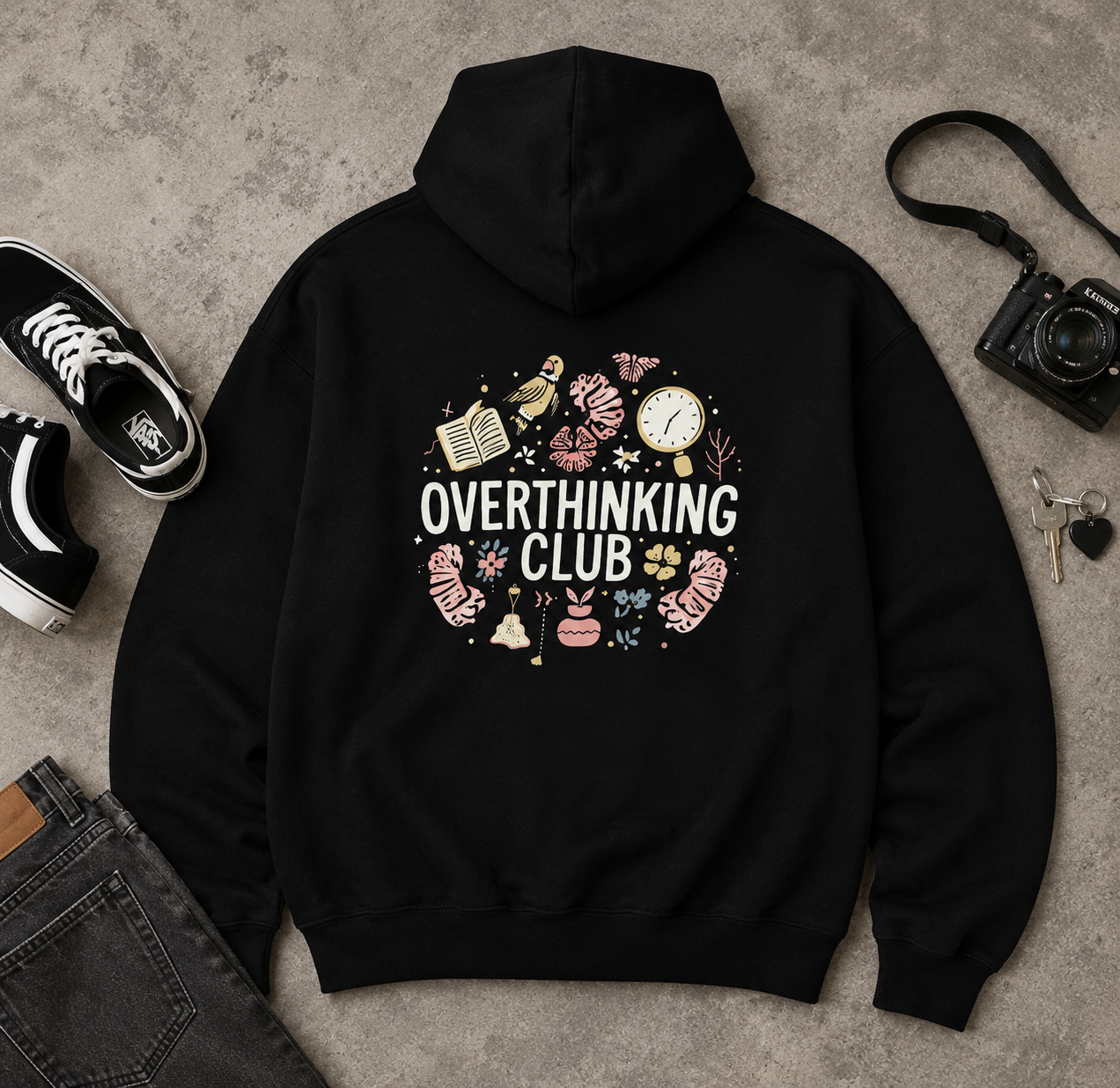 A black pullover hoodie laid flat on a textured gray surface, surrounded by styled props including black sneakers, a camera, keys, and part of a denim jacket. The hoodie has a large back graphic with the words 'OVERTHINKING CLUB' in white, surrounded by pastel doodles of books, a clock, brain imagery, and small icons.