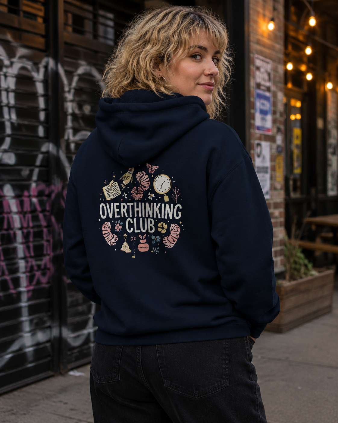 A woman wearing a navy blue hoodie with a circular graphic on the back that reads 'OVERTHINKING CLUB' surrounded by small illustrated objects, photographed outdoors in front of a storefront.