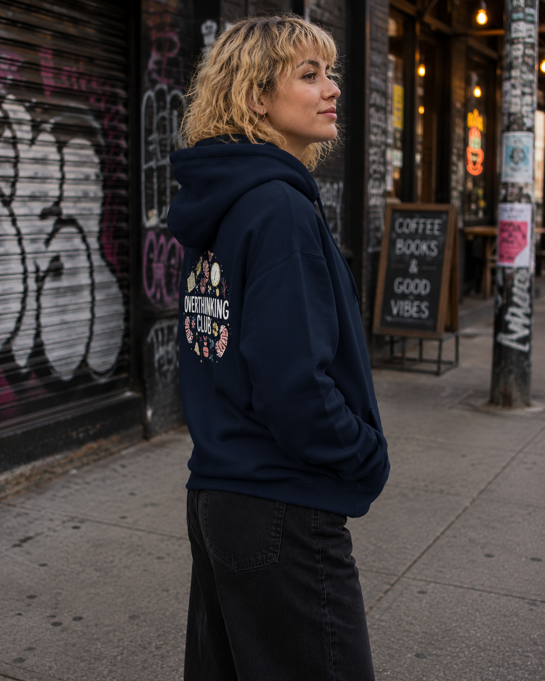 A woman with blonde hair stands on a city sidewalk wearing a navy blue hoodie with a circular graphic on the back that reads 'DETOURING CLUB' surrounded by small illustrations. The background shows a street scene with graffiti-covered walls, a storefront, and a coffee sign.