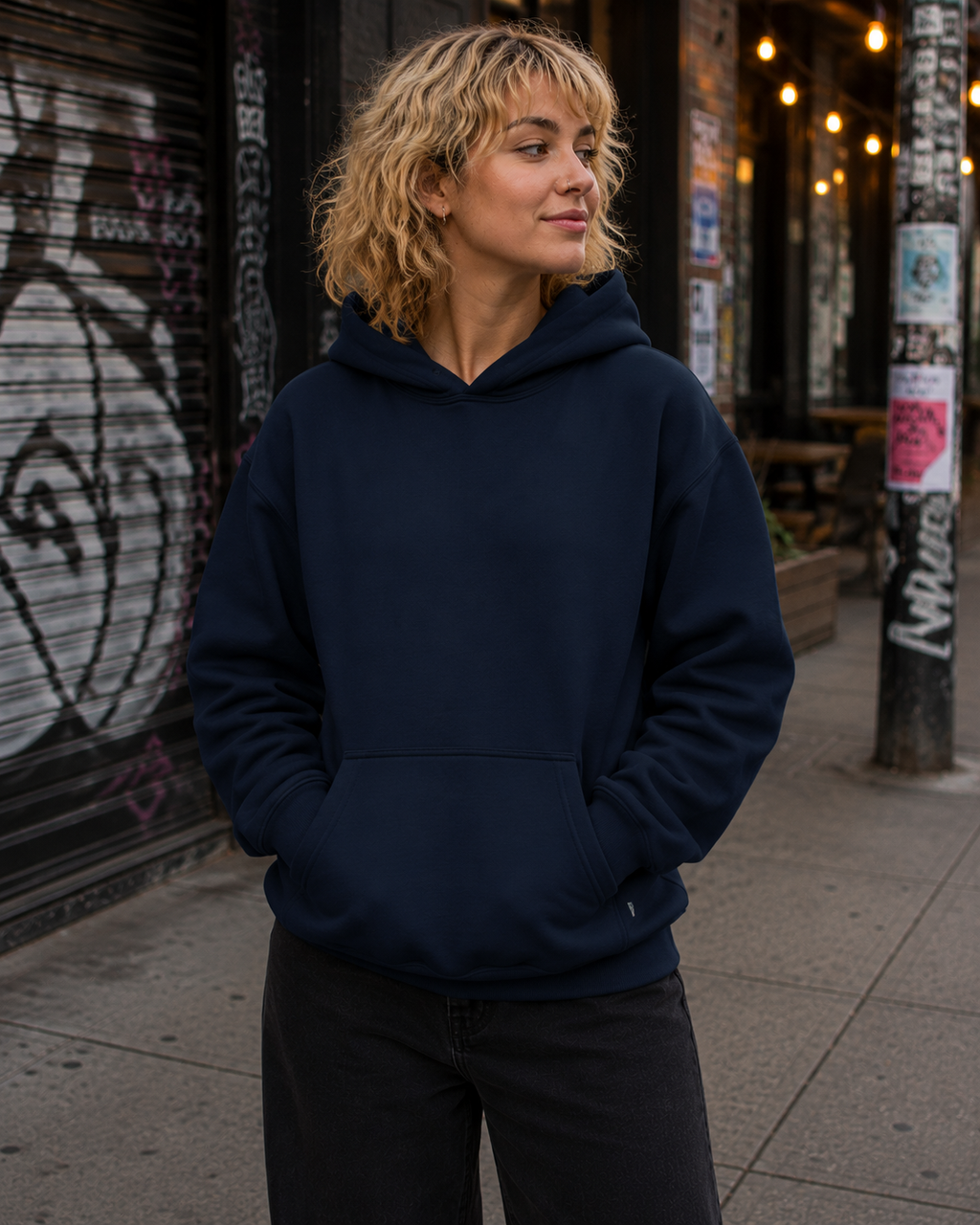 A woman with blonde curly hair wearing a navy blue pullover hoodie and black pants, standing on a city sidewalk in front of a graffiti-covered metal shutter.