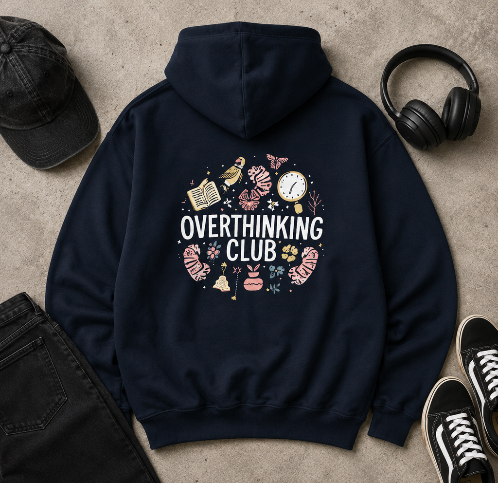 A navy blue hoodie laid flat on a textured concrete surface, surrounded by a black baseball cap, black over-ear headphones, black jeans, and black sneakers. The back of the hoodie features a large graphic with the words 'OVERTHINKING CLUB' in white, surrounded by small illustrated icons in pink, beige, and yellow.