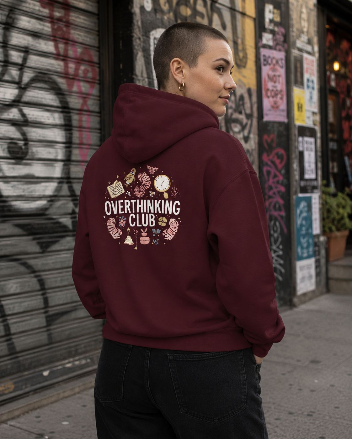 A person wearing a burgundy hoodie with a graphic on the back that reads 'OVERTHINKING CLUB' surrounded by illustrated objects, standing outdoors in front of a graffiti-covered wall.