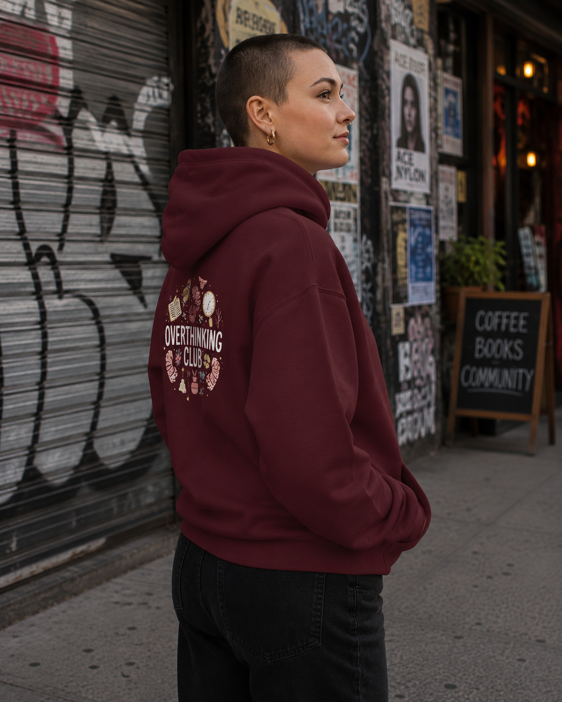 A person standing outdoors in front of a graffiti-covered metal shutter, wearing a burgundy hoodie with a circular graphic on the back that reads 'OVERTHINKING CLUB' and black jeans.