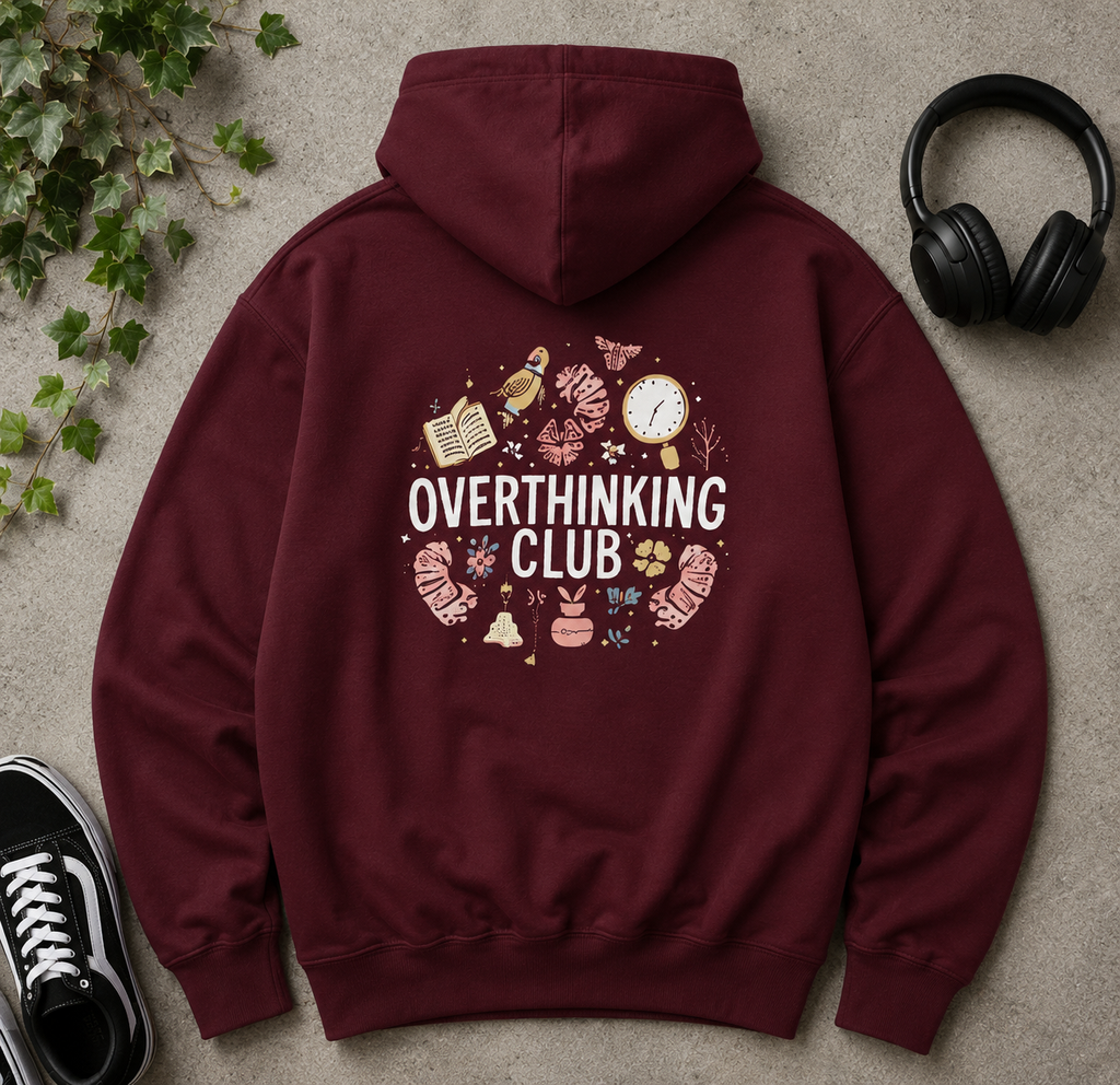 A maroon hoodie laid flat on a textured gray surface, surrounded by lifestyle props including black headphones, a black sneaker, and green ivy. The back of the hoodie features a circular graphic with the words 'OVERTHINKING CLUB' in white, surrounded by small illustrated objects in pink, beige, and brown tones.