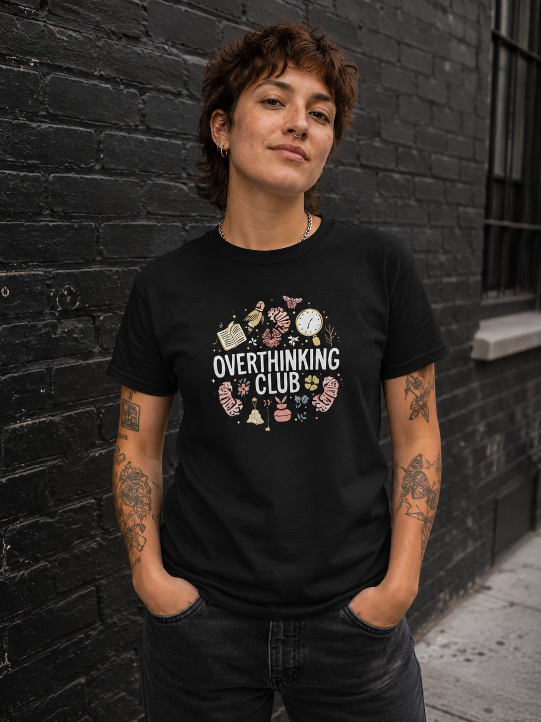 A person with short brown hair and tattoos stands against a dark brick wall wearing a black graphic T-shirt that reads 'OVERTHINKING CLUB' with small illustrated objects around the text, paired with dark jeans.