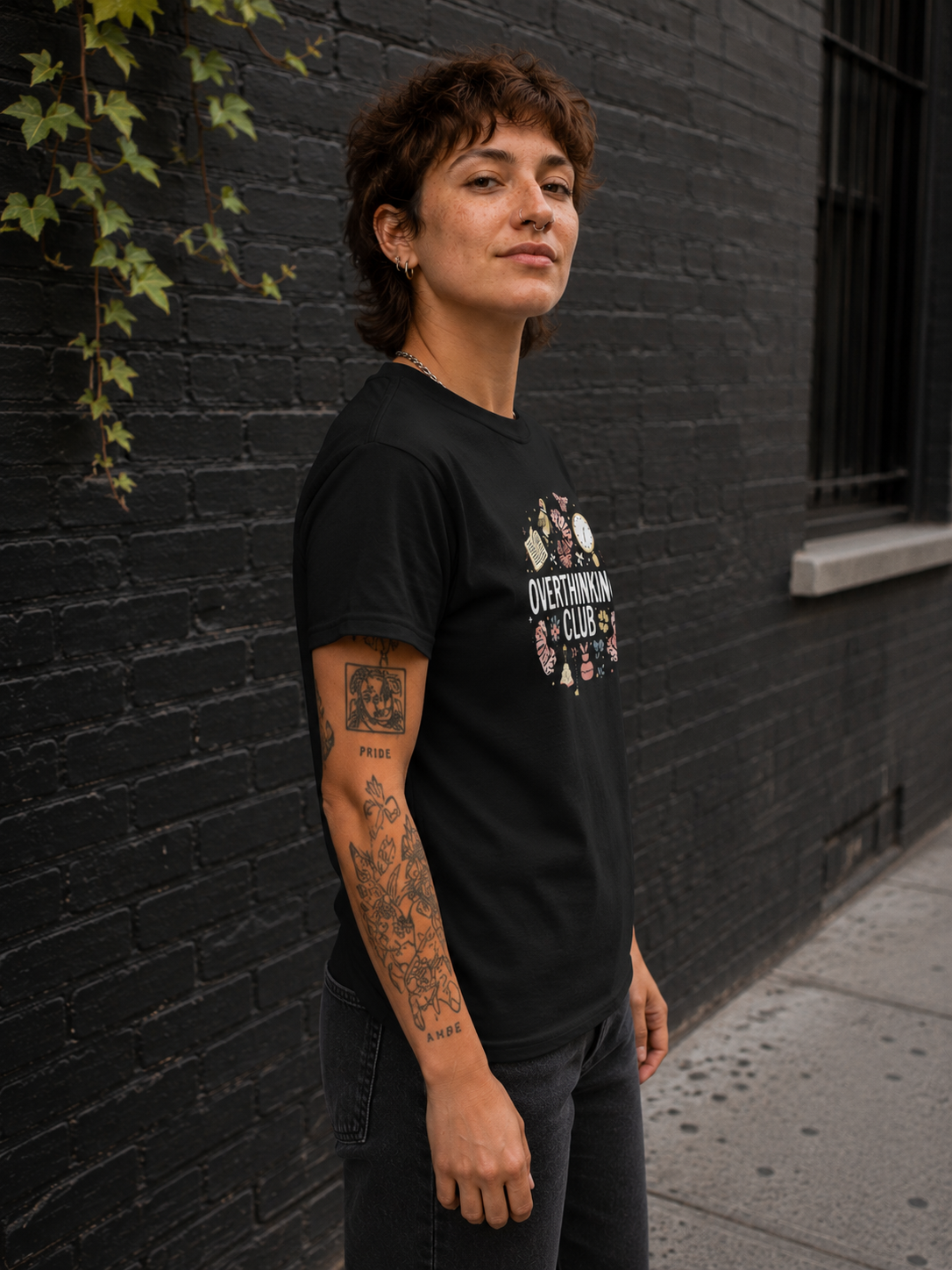 A person standing outdoors against a dark brick wall wearing a black graphic T-shirt with a white and pink 'Overthinking Club' print on the front.