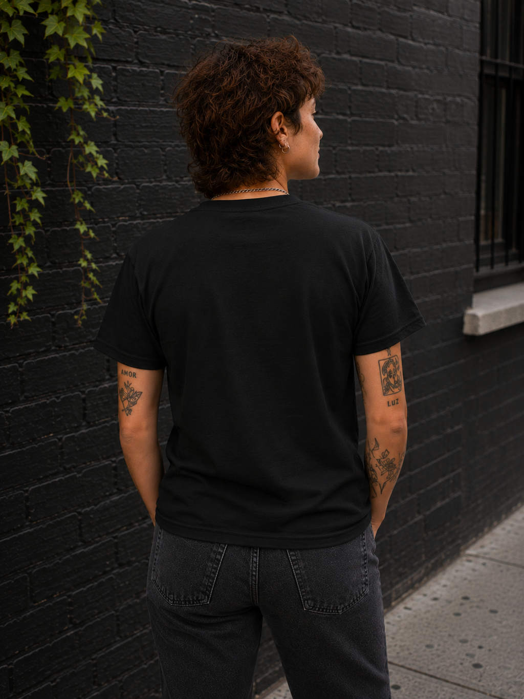 A person viewed from behind wearing a plain black short-sleeve T-shirt and dark gray jeans, standing outdoors against a black brick wall with ivy on the left.