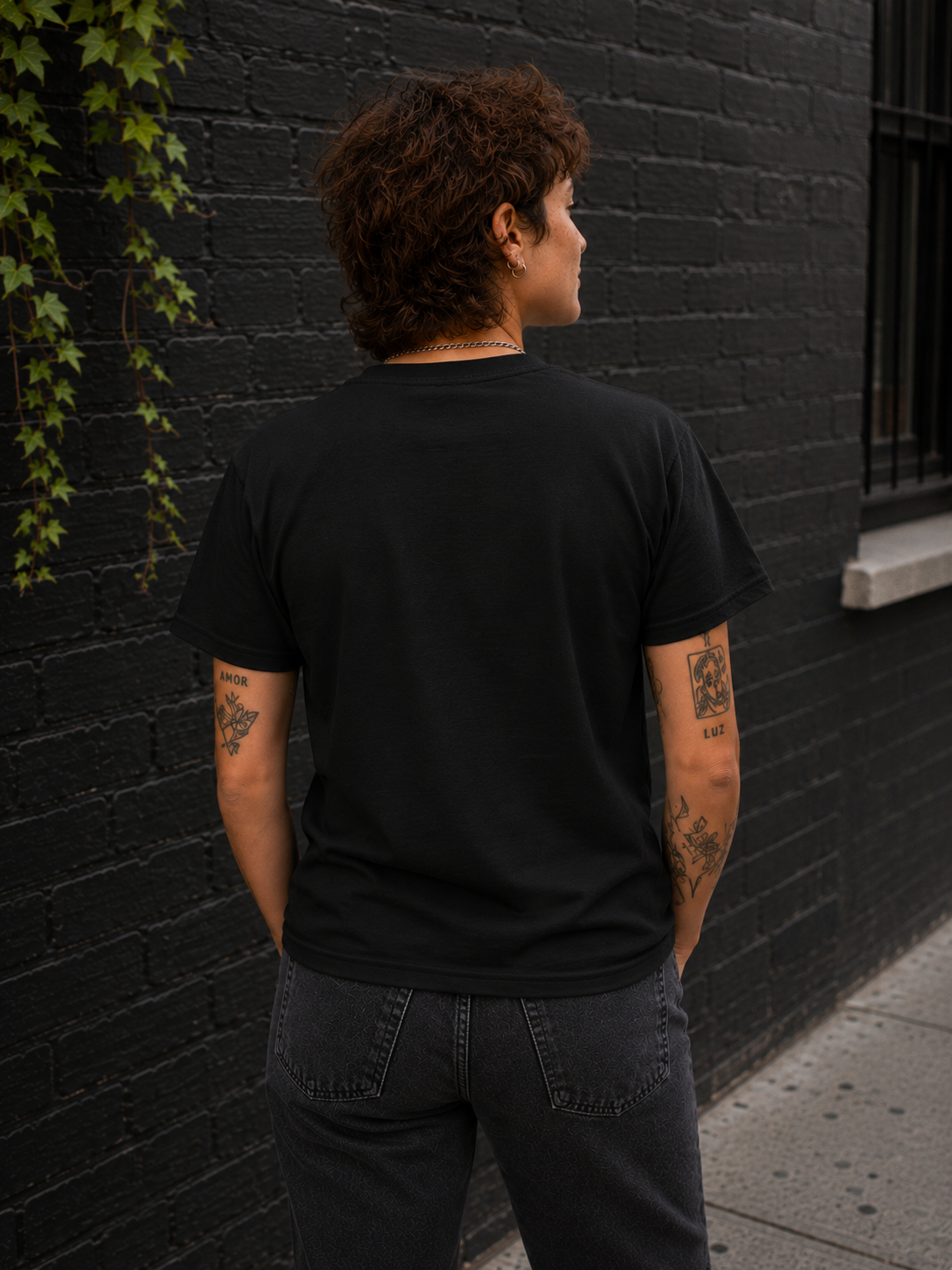 A person viewed from behind wearing a plain black short-sleeve T-shirt and dark gray jeans, standing outdoors against a black brick wall with ivy on the left.