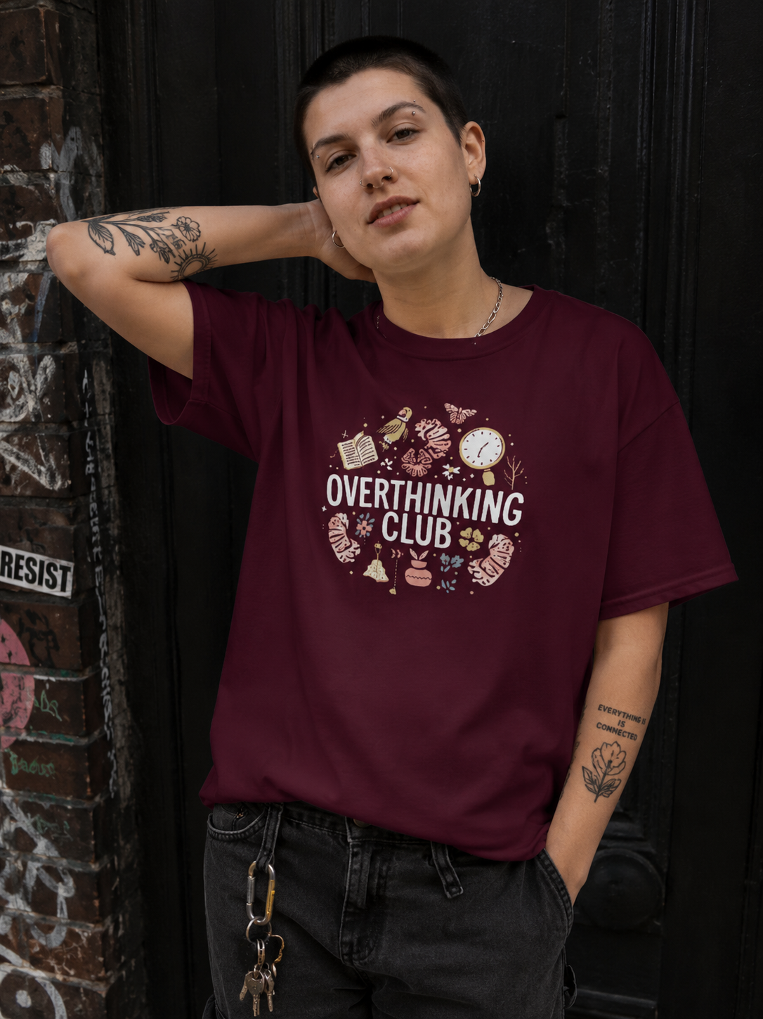 A person wearing a maroon short-sleeve graphic T-shirt with a circular 'Overthinking Club' print in cream and pink, standing against a dark wooden door and graffiti-covered brick wall.