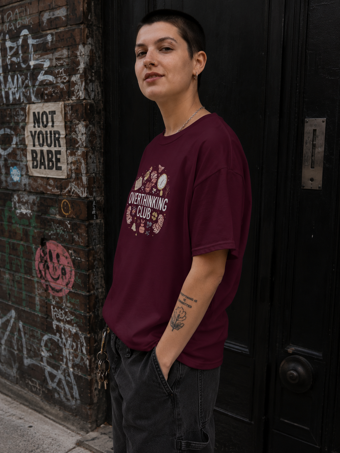 A person with a buzz cut stands in front of a graffiti-covered brick wall and a black door, wearing an oversized maroon t-shirt with a white graphic print that reads 'OVERTHINKING IS A CLUB'.