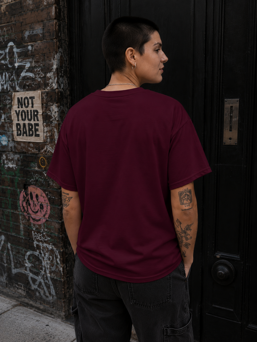 A person with short dark hair stands in front of a black door and graffiti-covered wall, wearing an oversized maroon short-sleeve T-shirt and dark cargo pants, shown from the back.