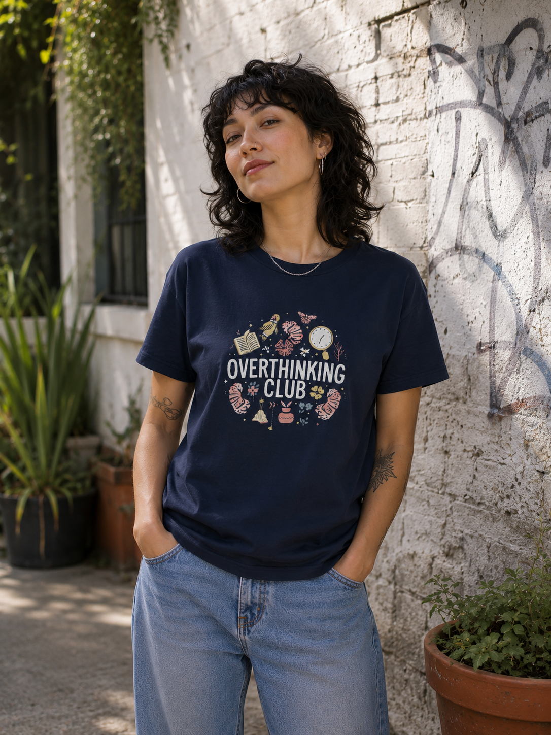 A woman wearing a navy blue graphic t-shirt with the text 'OVERTHINKING IS A CLUB' and small illustrations, paired with light blue jeans, standing against a white brick wall.