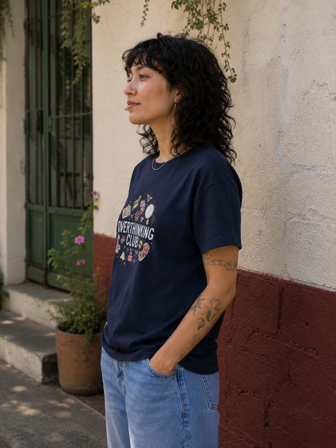 A woman with curly black hair stands outdoors against a textured cream and terracotta wall, wearing a navy blue graphic t-shirt with the text 'OVERTHROWING CLUB' and light blue jeans.