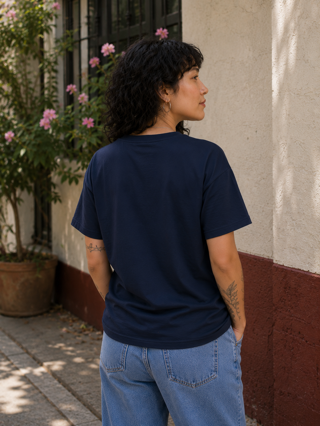 A woman with curly dark hair is shown from behind wearing a navy blue short-sleeve crewneck t-shirt and light blue jeans, standing outdoors next to a textured beige and reddish-brown wall with potted pink flowers.