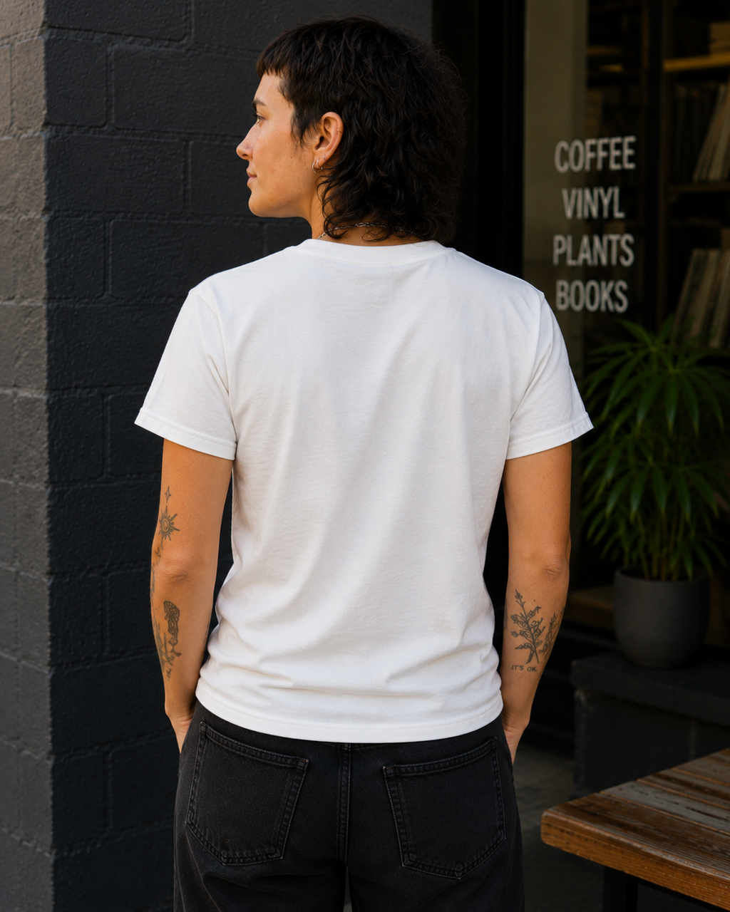 A person standing outdoors in front of a dark brick wall and storefront window, wearing a plain white short-sleeve T-shirt and dark jeans. The shirt is shown from the back, with visible tattoos on both arms.