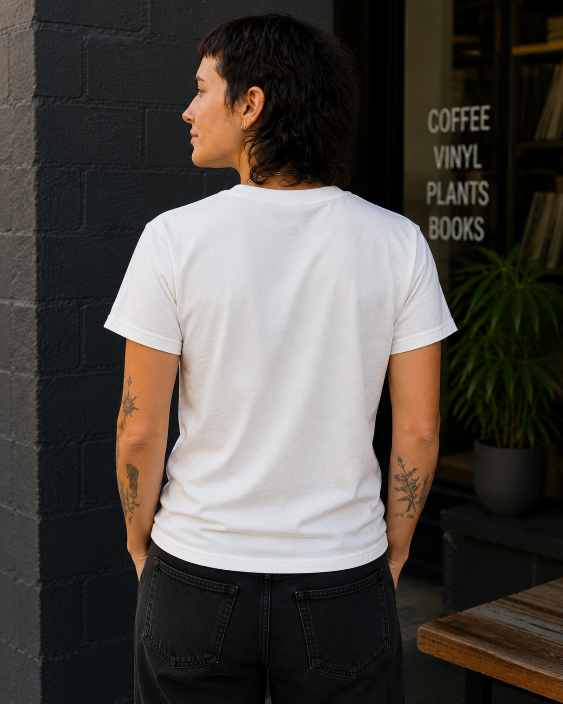 A person standing outdoors in front of a dark brick wall and storefront window, wearing a plain white short-sleeve T-shirt and dark jeans. The shirt is shown from the back, with visible tattoos on both arms.