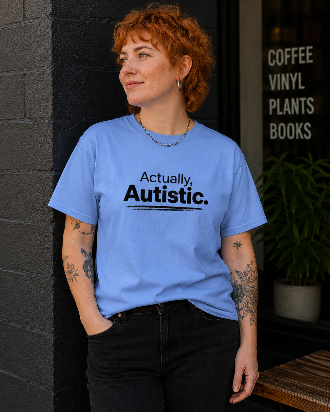 A woman with short red hair stands outdoors wearing a light blue graphic T-shirt that reads 'Actually, Autistic.' in black text, paired with black pants.