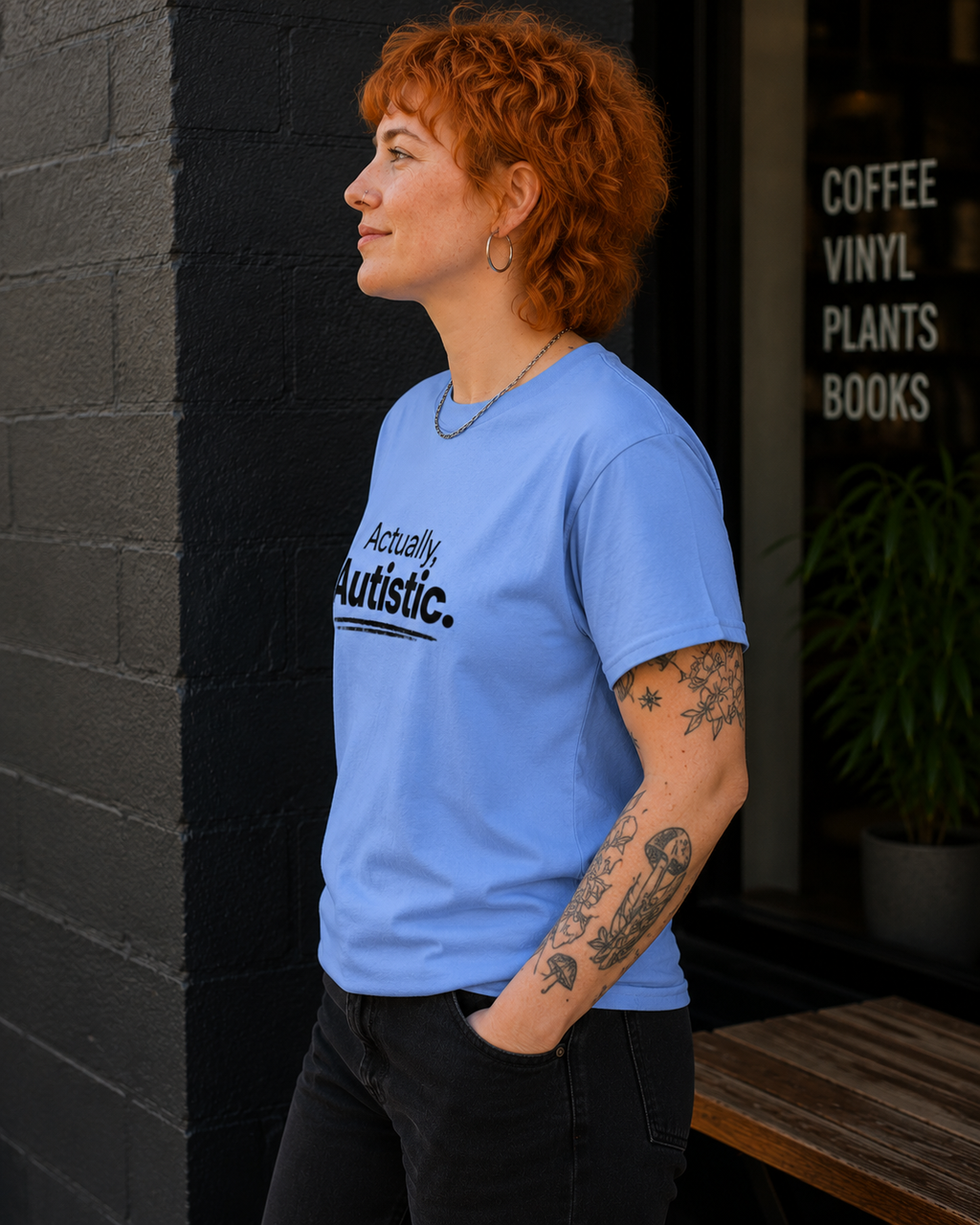 A woman with short curly red hair stands in profile wearing a light blue graphic T-shirt that reads 'Actually Autistic.' in black text.