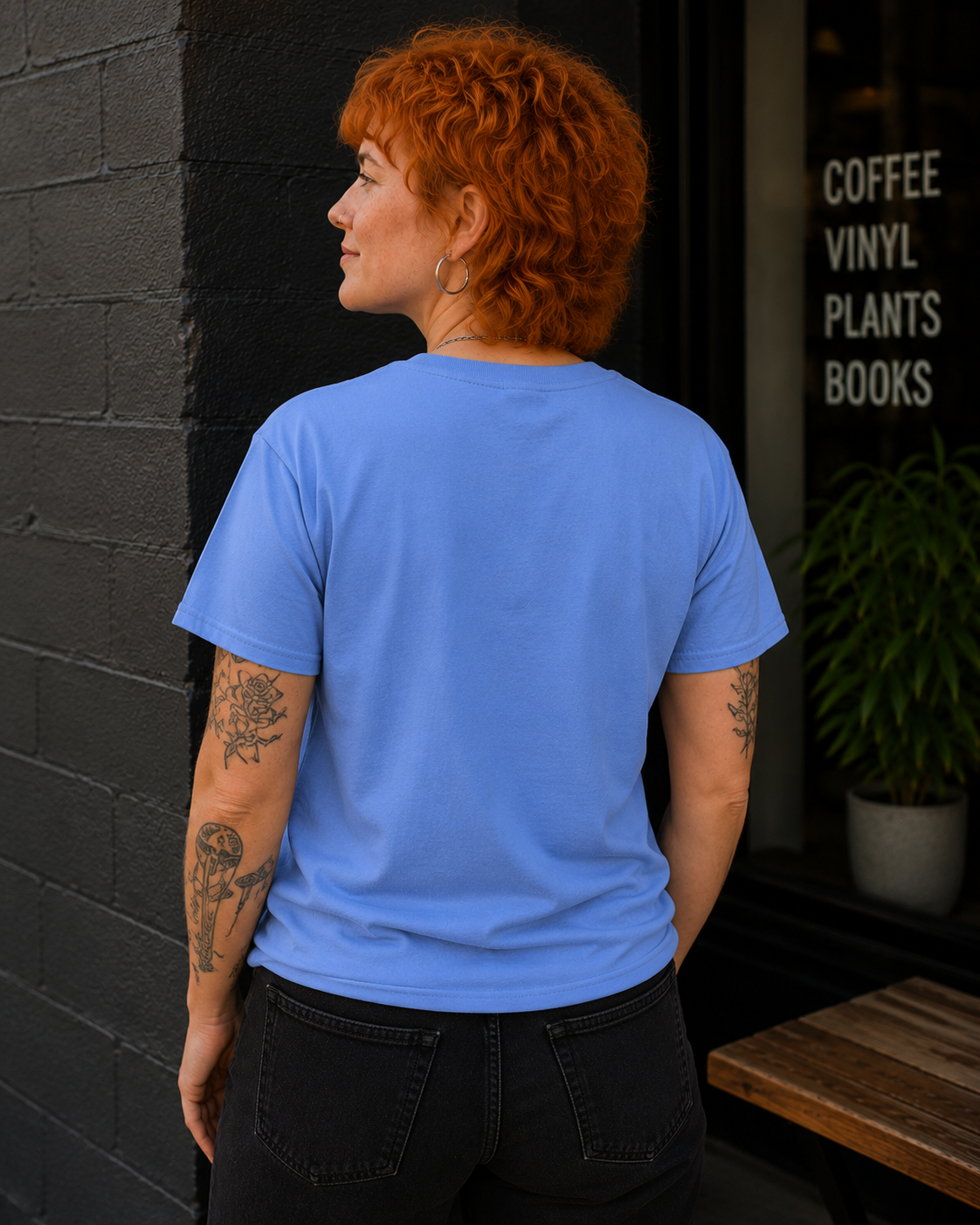 A person with short curly red hair stands with their back to the camera wearing a light blue crewneck t-shirt and black jeans, standing next to a dark brick wall and a storefront window with text that reads 'COFFEE VINYL PLANTS BOOKS'.